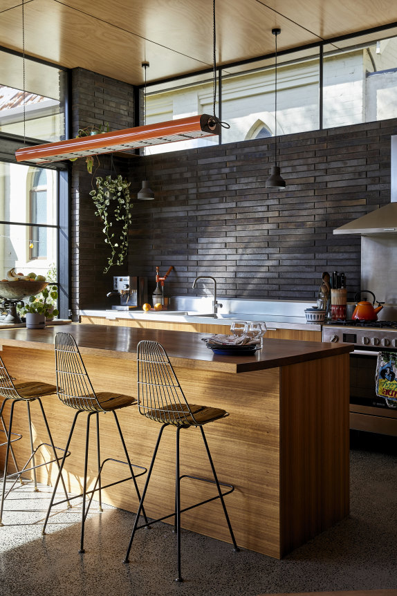 L'îlot de cuisine est fait de bois noir et comprend des chaises de bar rétro Framac. Derrière se trouvent un plan de travail en acier inoxydable et un grand four et une cuisinière Bosch.