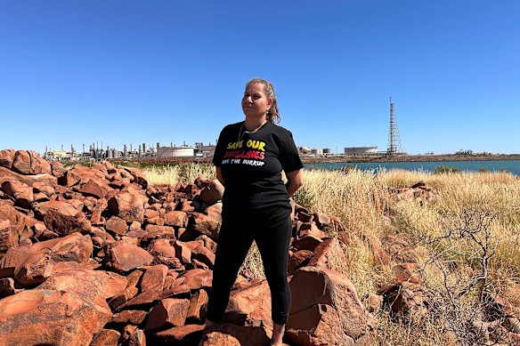 Murujuga Gustodian Raelene Cooper se tenant sur Murujuga, avec l'usine de Karratha de Woodside en arrière-plan.
