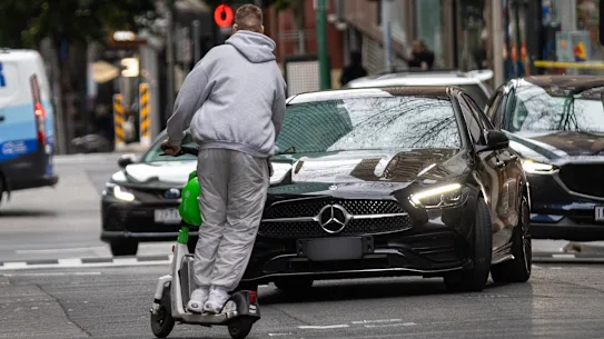 Un homme conduit un scooter électrique dans le mauvais sens dans Little Bourke Street sans casque en 2024, faisant un écart pour rater une voiture venant en sens inverse. 