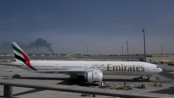 Un panache de fumée provoqué par une frappe iranienne est visible en arrière-plan, tandis qu'un avion d'Emirates est stationné à l'aéroport international de Dubaï.